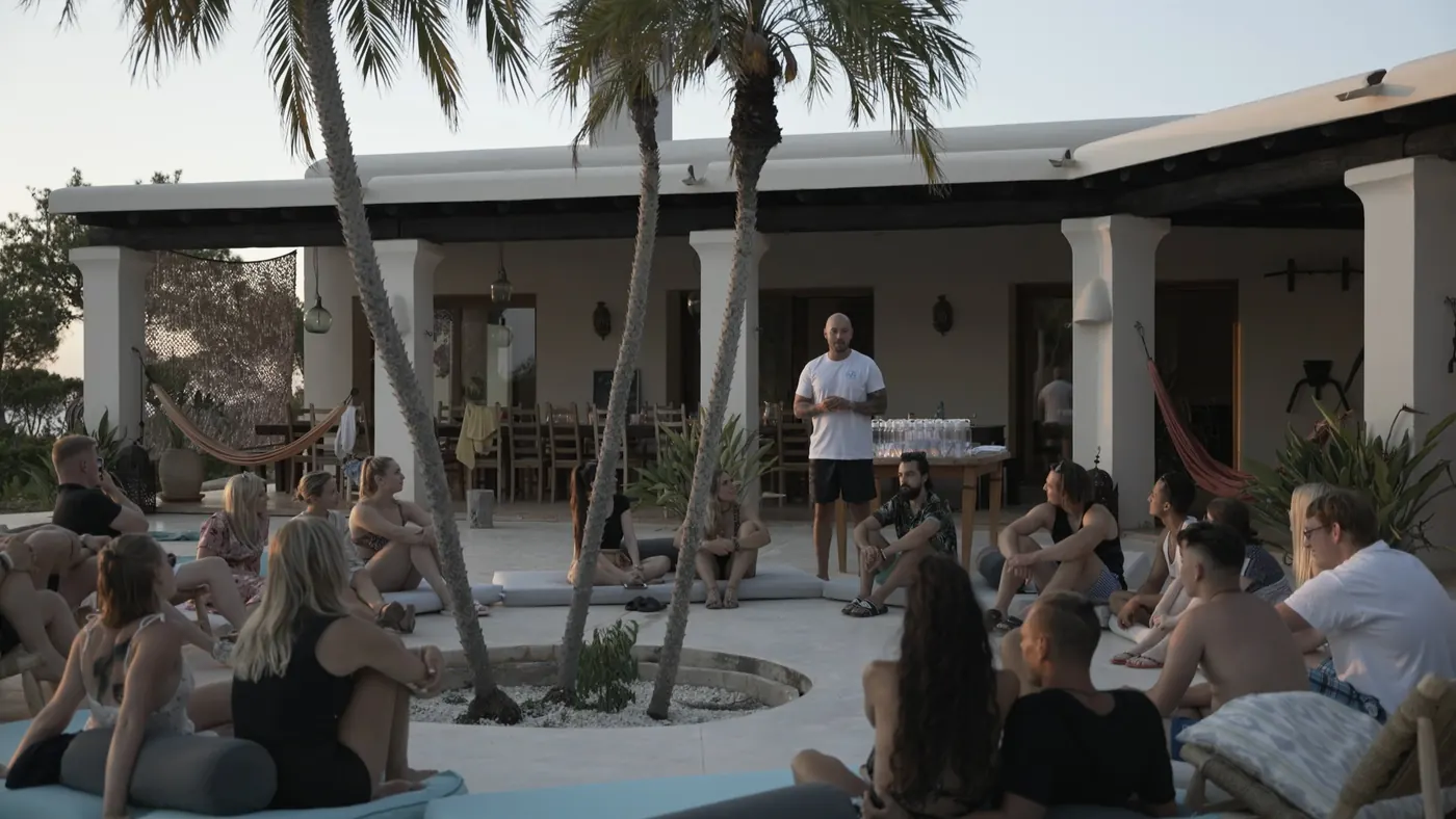 Adrian spricht vor einer Gruppe im Kreis auf einer Terrasse unter Palmen